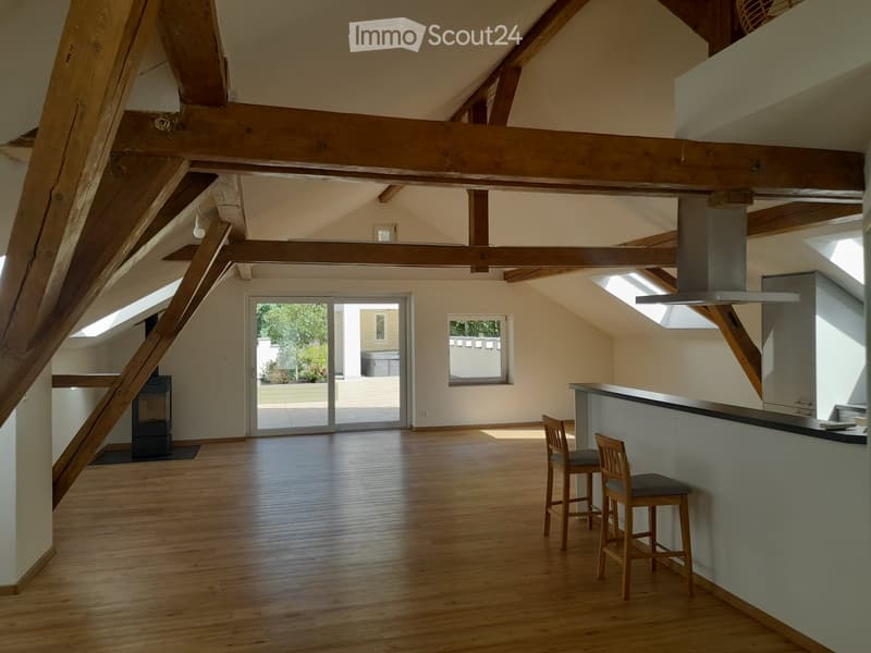 Bright, open loft-style room with wooden beams, hardwood floors, and large windows. Features a kitchen island with two stools, a fireplace, and a sliding glass door leading to a patio.