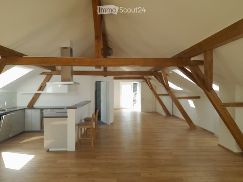 Open-plan attic space with exposed wooden beams, wooden flooring, and a modern kitchen area featuring a white island, stools, and stainless steel appliances.