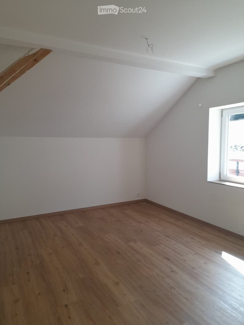 Empty room with wooden flooring, white walls, and a slanted ceiling. A window with a white frame is visible on the right side.