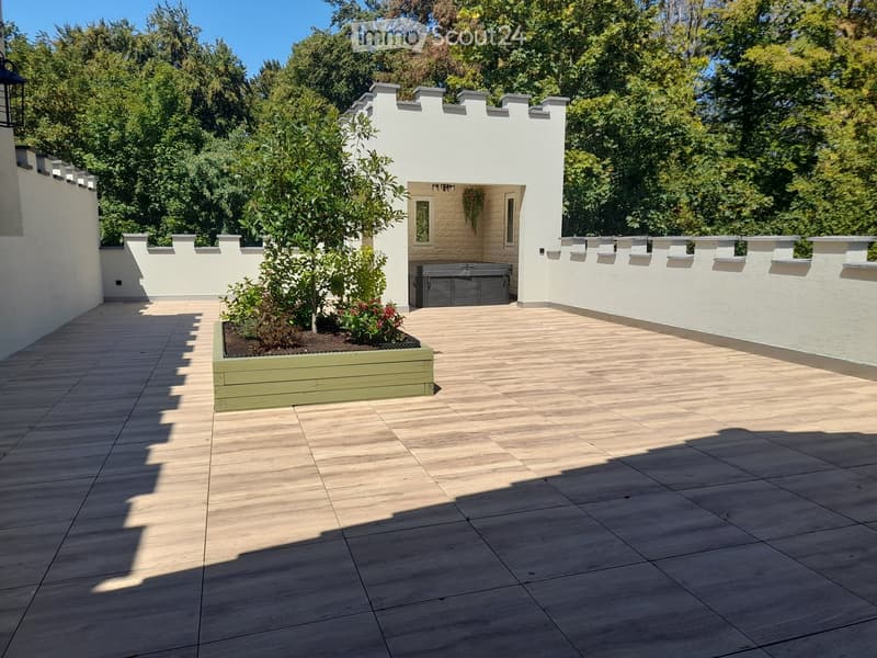 Wooden deck patio with raised planter box, small seating area with built-in bench, surrounded by white walls and greenery.