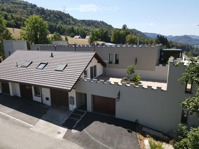 Modern two-story house with a tiled roof, garage, and a small balcony area with plants. The house is surrounded by a scenic, green landscape with hills in the background.