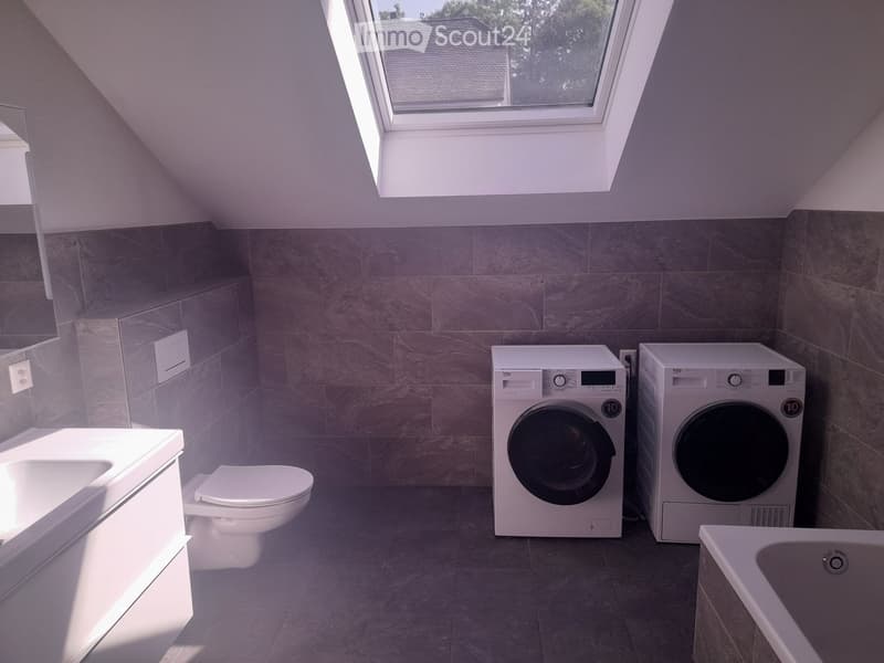 Modern bathroom with a toilet, bathtub, and two stacked washing machines/dryers, featuring large windows and sleek tilework.