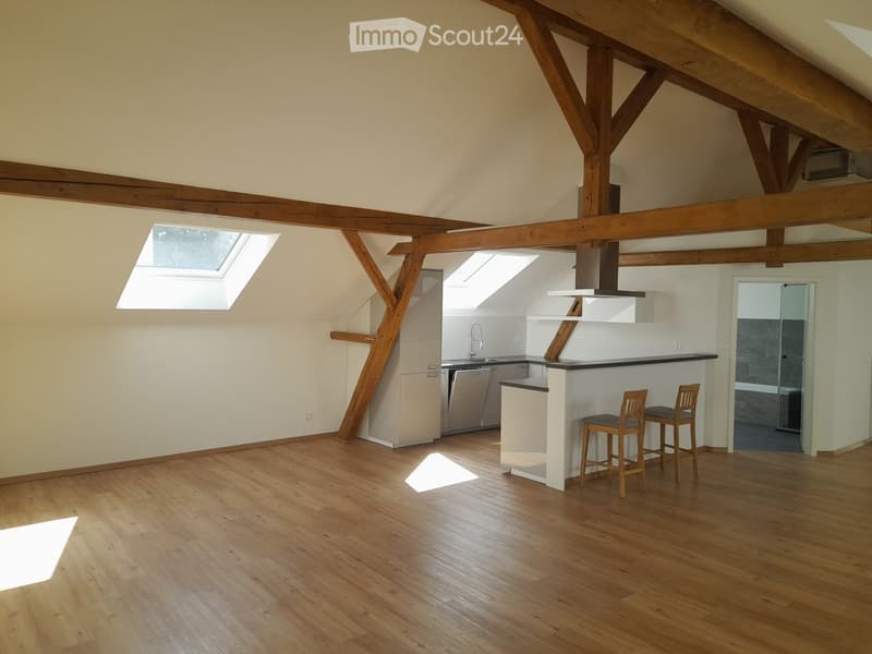 Open-plan room with wooden beams, wooden flooring, kitchen area with island, two stools, dishwasher, and a small bathroom area visible.