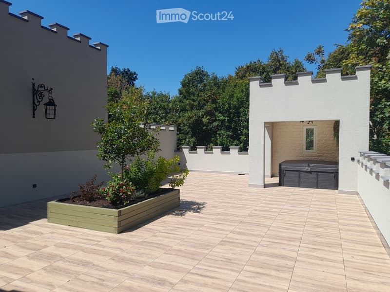 A modern outdoor patio with wooden decking, a raised planter box with plants, a hot tub, and surrounding white walls with decorative battlements.