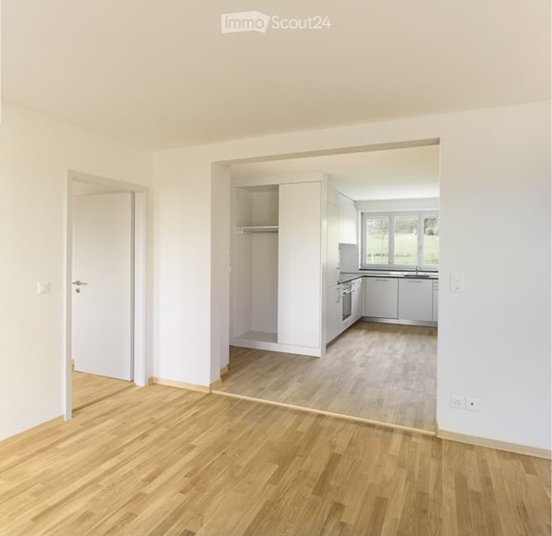 Empty room with wooden flooring and white walls, featuring an open doorway leading to a modern kitchen area with white cabinets and stainless steel appliances.