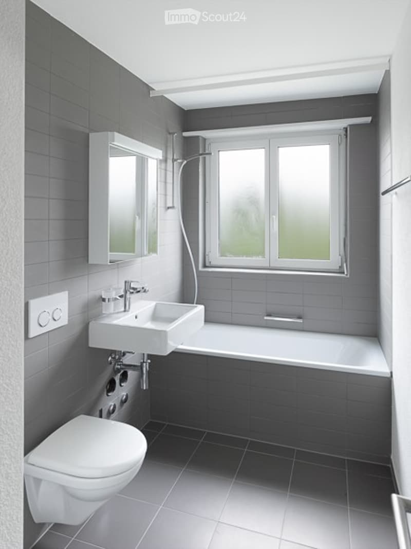 Modern bathroom with grey tiled walls and floor, white fixtures including a sink, toilet, and bathtub, a large window, and a mirror cabinet.