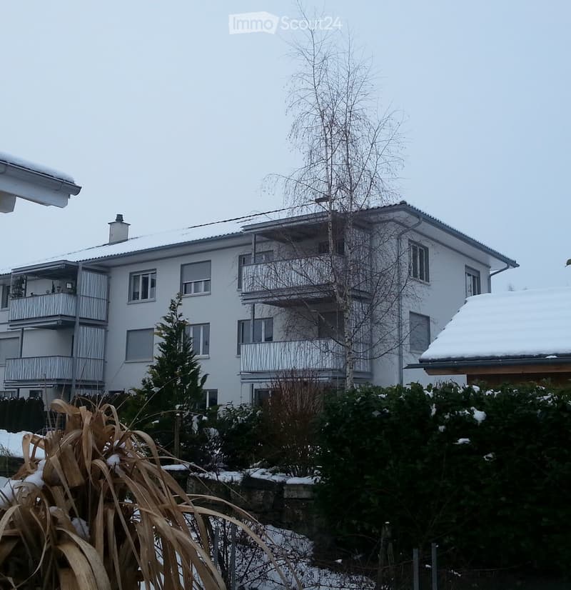 A two-story white building with multiple windows and balconies, surrounded by snow-covered trees and bushes.