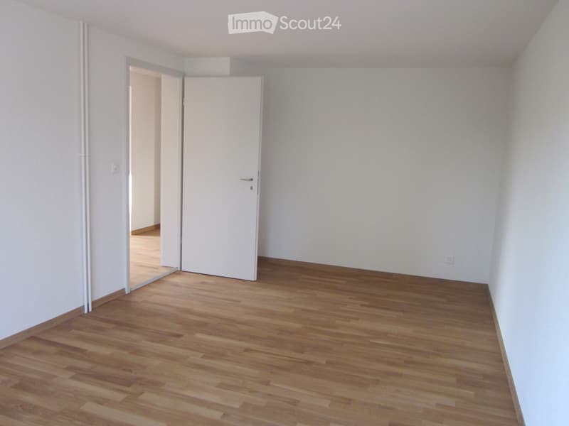 Empty room with light wooden flooring and white walls, featuring a closed white door and an open doorway leading to another space.