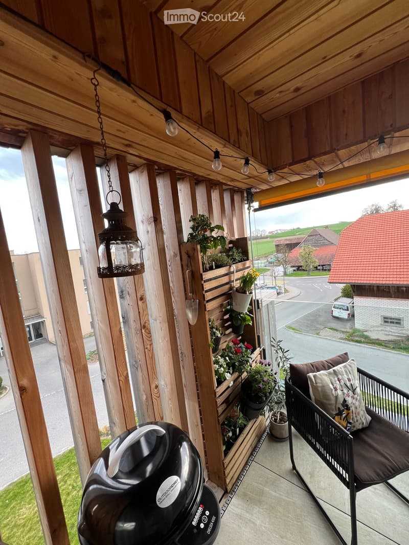 Wooden balcony with a hanging lantern, potted plants, a black coffee maker, and a cushioned chair with a decorative pillow.