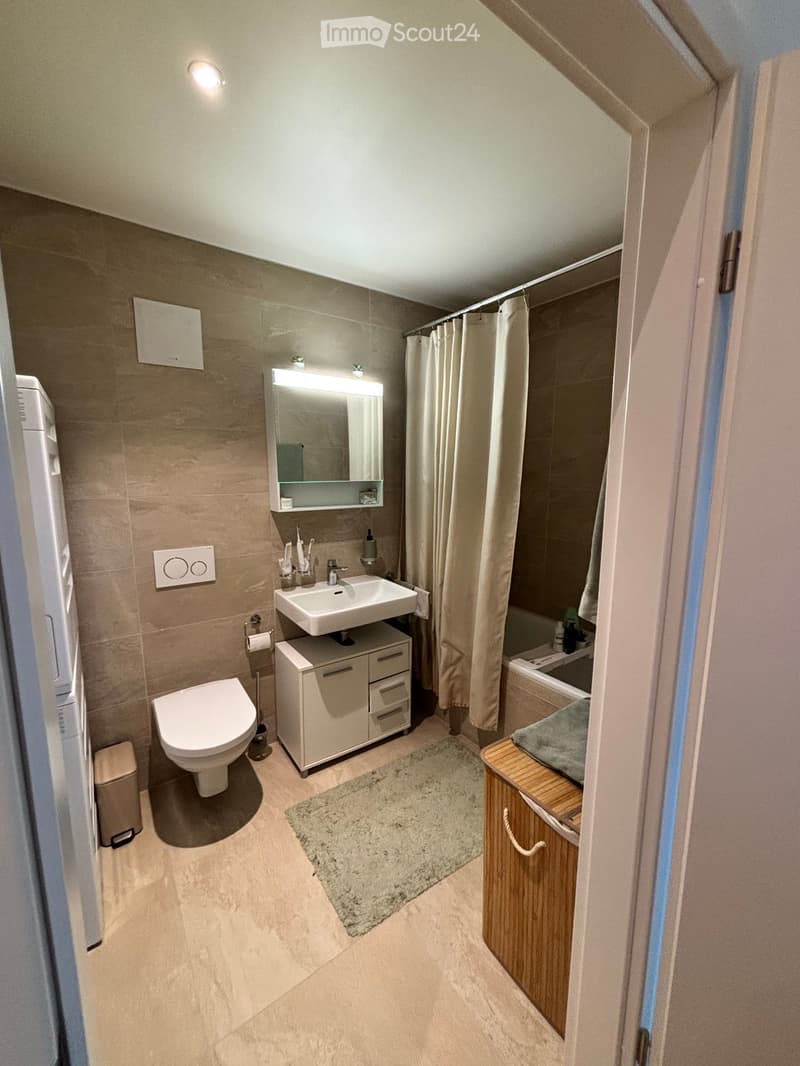 Modern bathroom with beige tiles, a toilet, a sink with a mirrored cabinet, a bathtub with a shower curtain, and a small wooden shelf.
