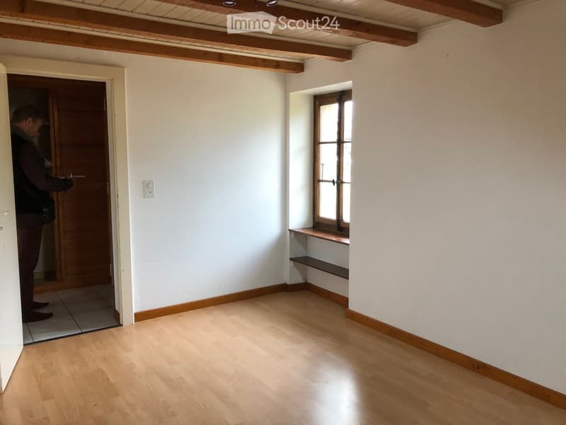 Empty room with wooden flooring, white walls, and a window with wooden frame. There is a small wooden bench under the window and a door leading to another room.