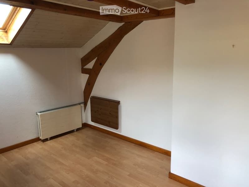 Empty room with wooden flooring, white walls, exposed wooden beams on the ceiling, and a skylight window. Radiators are visible on the walls.