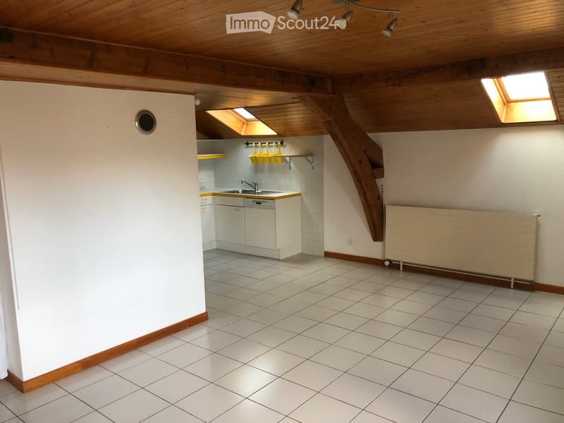 The image shows a small, well-lit kitchen area with white tiled flooring, white cabinets, a sink, a dishwasher, and a radiator. A wooden staircase leads upwards, and the ceiling features exposed wooden beams and recessed lighting.