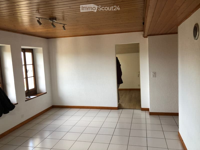 Empty room with white tiled floor, white walls, wooden trim, and a wooden ceiling with track lighting. A window with wooden frame and a small shelf is visible on one wall, and an open doorway leads to another room.