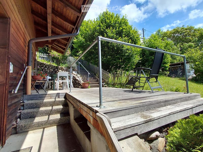 Wooden deck with metal railing, steps leading up, outdoor furniture including a chair and table, surrounded by greenery and a stone wall.