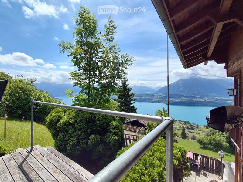Balcony with wooden decking, metal railing, overlooking a scenic lake and mountain landscape with trees and distant houses.