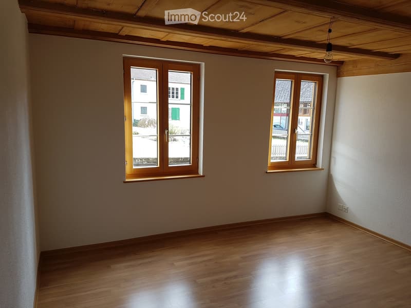Empty room with wooden flooring, exposed wooden ceiling beams, and two windows with wooden frames. The walls are painted in a light neutral color.