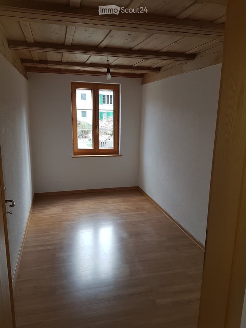 Empty room with wooden flooring, white walls, and a large window with wooden frame. Exposed wooden ceiling beams and a single hanging light fixture are visible.