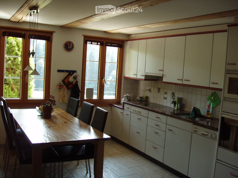 Bright kitchen with white cabinets, wooden dining table, and six chairs. Large windows provide natural light, and the space includes an oven, dishwasher, and sink.