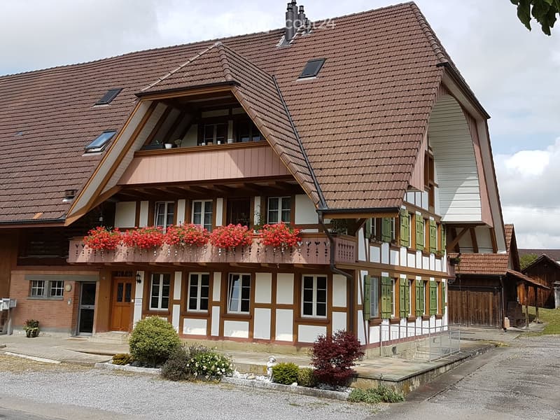 Two-story traditional house with red-tiled roof, white and brown facade, balcony with red flowers, green shutters, and small garden area.