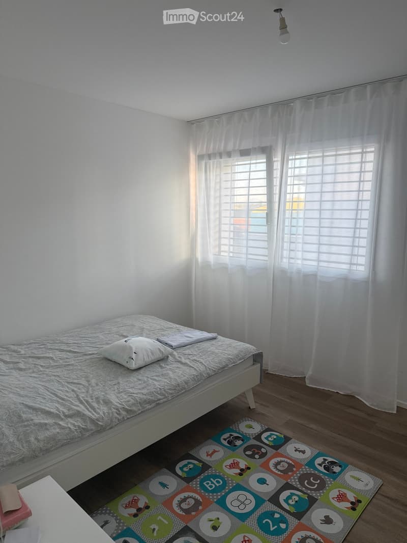 Minimalist bedroom with a single bed, white pillow, and a colorful rug on the floor. The room features white walls, wooden flooring, and large windows with white curtains.