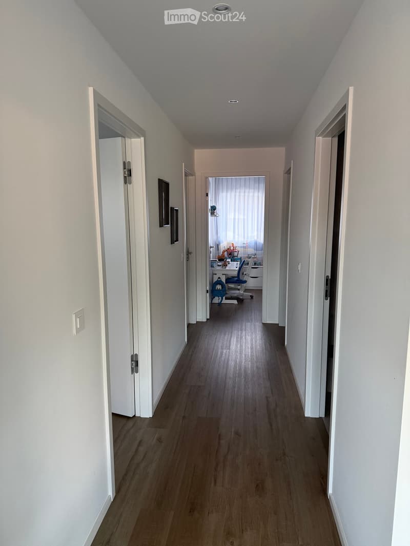 A long, narrow hallway with light-colored walls and wooden flooring, featuring several open doors leading to adjacent rooms.