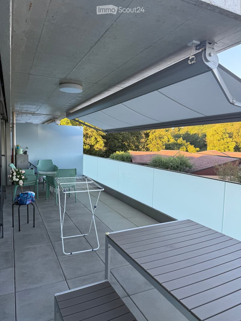 Modern balcony with grey wooden bench, white metal table, green chairs, and a view of trees and rooftops.