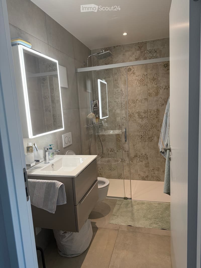 Modern bathroom with a white sink, mirror with LED lighting, glass-enclosed shower, and a toilet. Beige tiles and a light-colored floor.