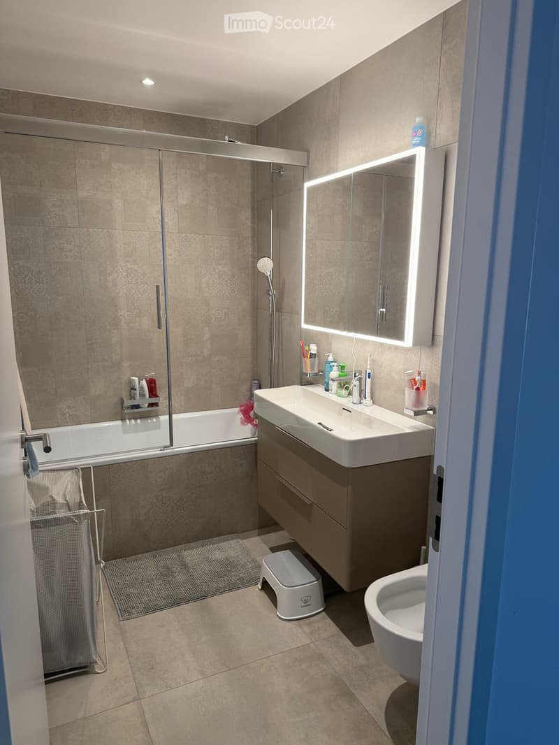 Modern bathroom with a bathtub, sink, toilet, and a large illuminated mirror above the sink. The space features beige tiles, a towel rack, and a small stool near the toilet.