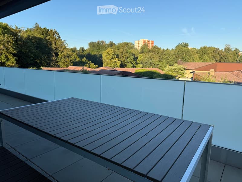 Modern balcony with a wooden table, glass railing, and a view of trees and buildings.