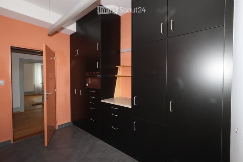 An empty kitchen with open door, black cabinets, marble counter, wooden shelves, orange walls, hardwood floors