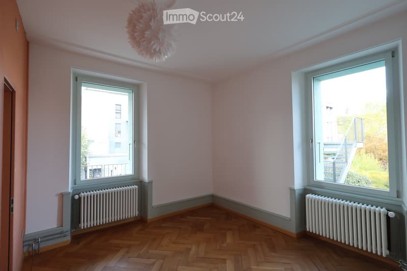 Empty room, white walls, white ceiling, white radiators, two windows, wooden parquet flooring, view of stairs from one window