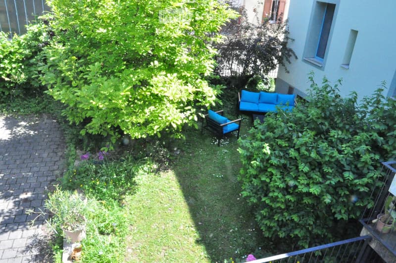 A small, lush garden with a brick pathway, green grass, and a seating area featuring a blue sofa and matching chair.