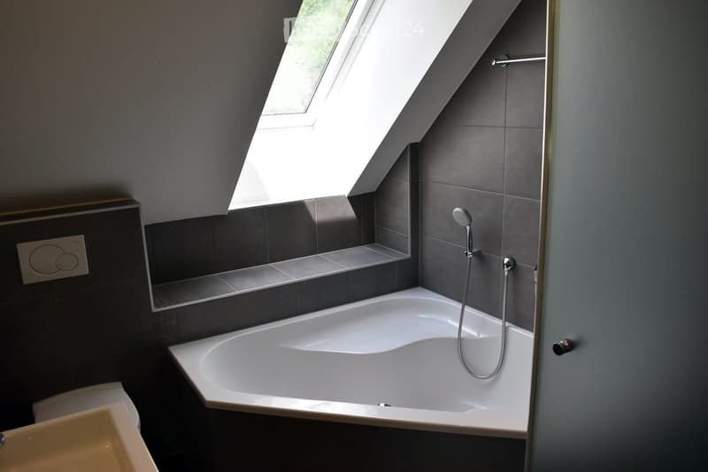 Modern bathroom with a white bathtub, showerhead, sink, mirror, and built-in shelf. The room features dark grey tiles and a skylight window providing natural light.