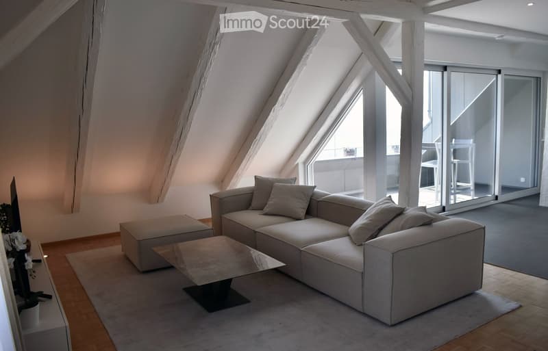 Modern living room with a grey sectional sofa, coffee table, ottoman, and a chair near a large window with a view of a balcony.