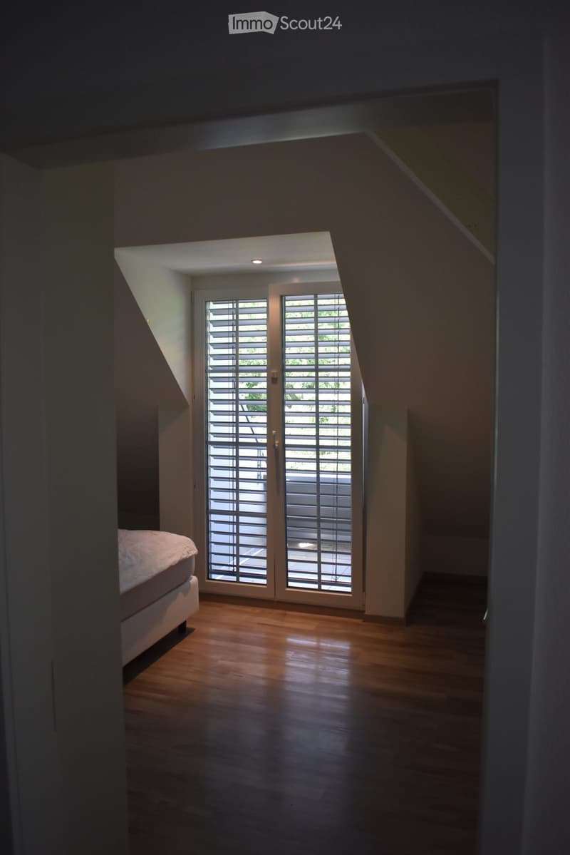 Bedroom with wooden flooring, a bed on the left, and large sliding glass doors with blinds leading to an outdoor area.