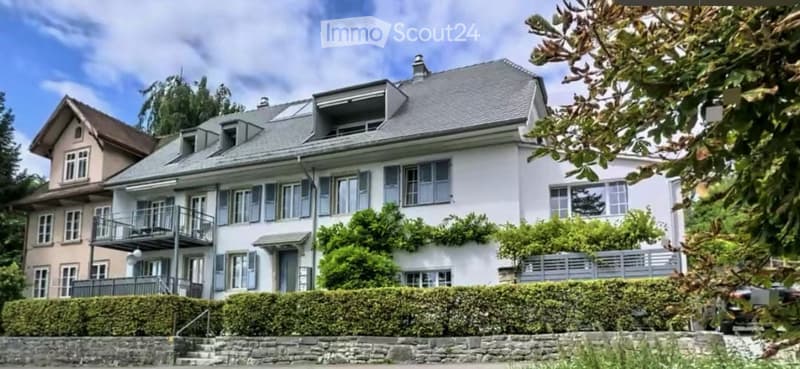 A multi-story residential house with a grey roof, multiple windows, and a balcony on the upper floor. The house is surrounded by greenery and has a stone pathway leading to the entrance.