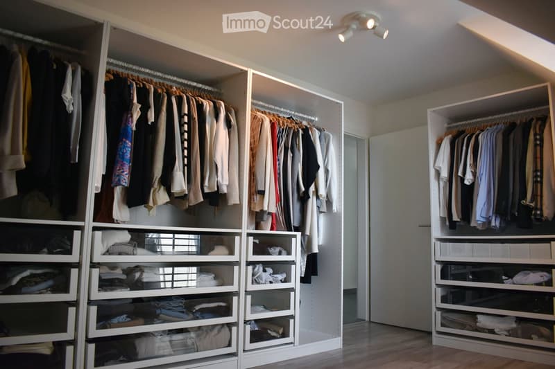 A spacious, well-organized walk-in closet with white built-in shelving units, multiple hanging rods, and neatly arranged clothing and folded items. The room features wooden flooring and recessed lighting.