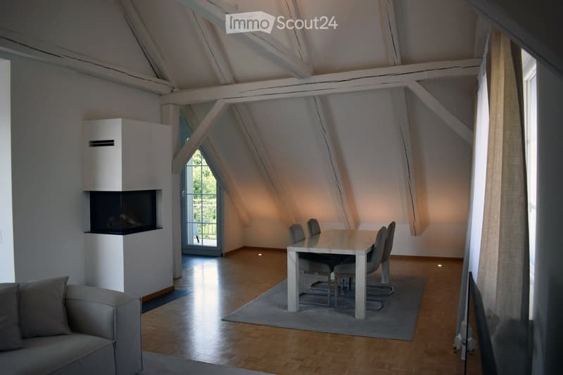 Bright, open loft-style room with wooden flooring, exposed wooden beams, a grey sofa, a dining table with chairs, and a modern fireplace.
