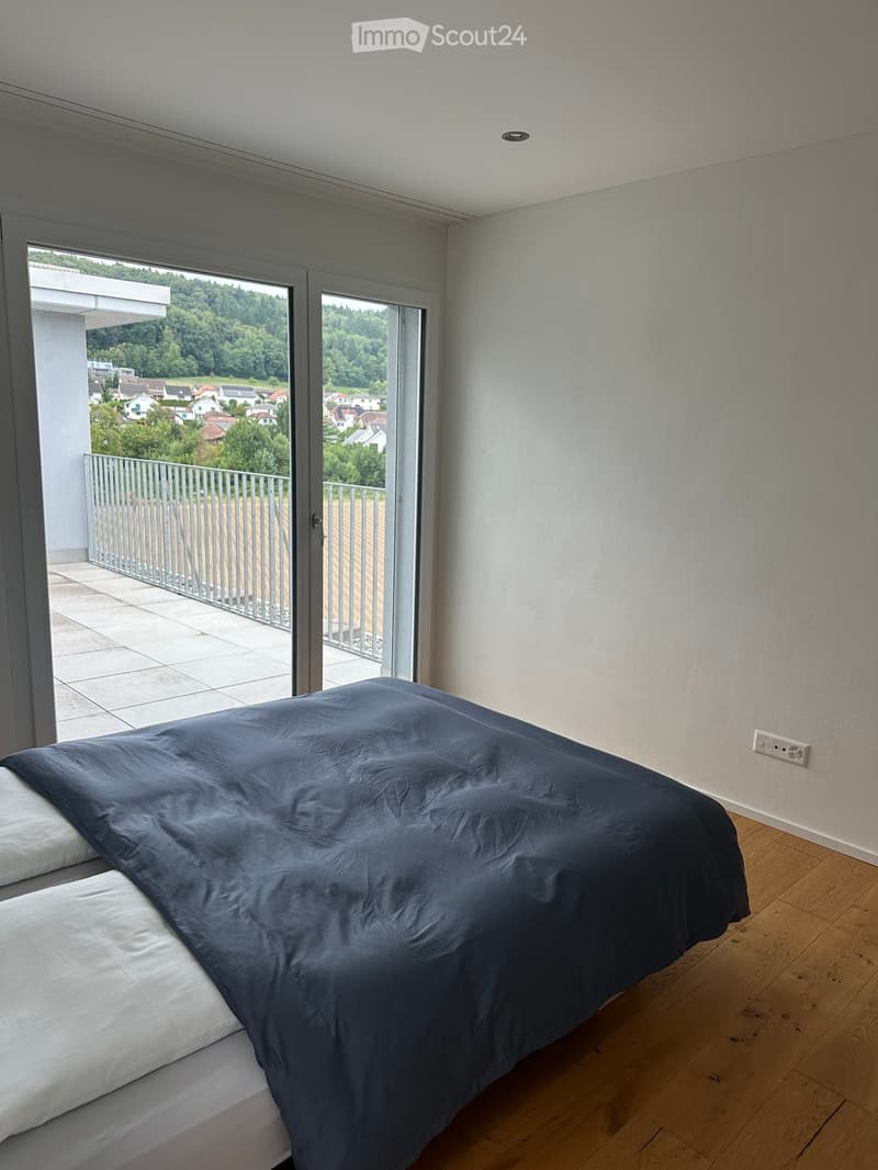 Modern bedroom with wooden flooring, large window opening to a balcony with a scenic view, and a neatly made bed with dark blue bedding.