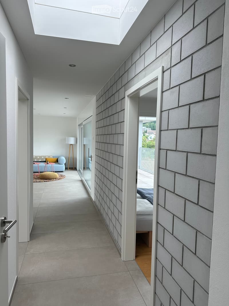 Modern hallway with grey tiled floor, white walls, and a skylight ceiling. Features a glass door leading to a balcony and a partially visible living area with a sofa and floor lamp.