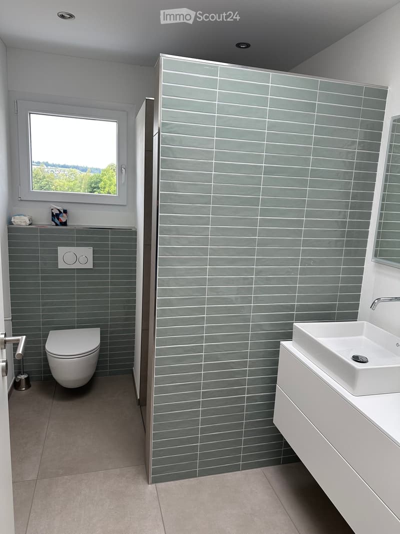 Modern bathroom with grey tiled walls, white toilet, white sink, and a large window offering a view of greenery.
