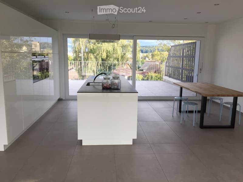 Modern kitchen with white island, sink, wooden dining table, and four stools, overlooking a balcony with outdoor view.
