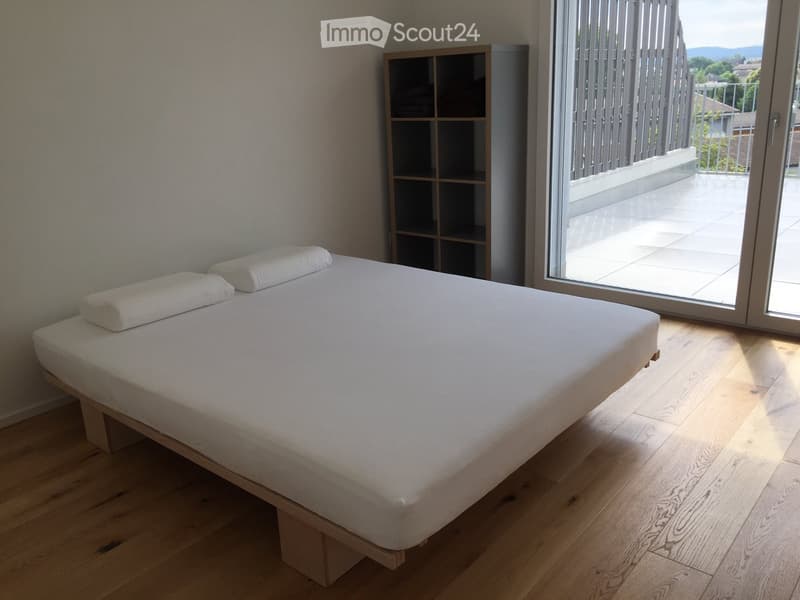 Minimalist bedroom with a single bed, white linens, and a small shelving unit near a large sliding glass door leading to a balcony.