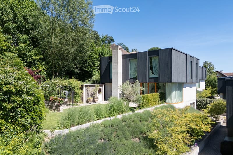 Modern two-story house with dark wooden facade and large windows, surrounded by a well-maintained garden with various plants and trees.