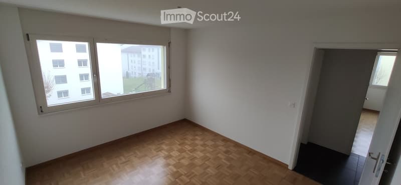 Empty room with wooden flooring, white walls, and large windows offering a view of neighboring buildings.