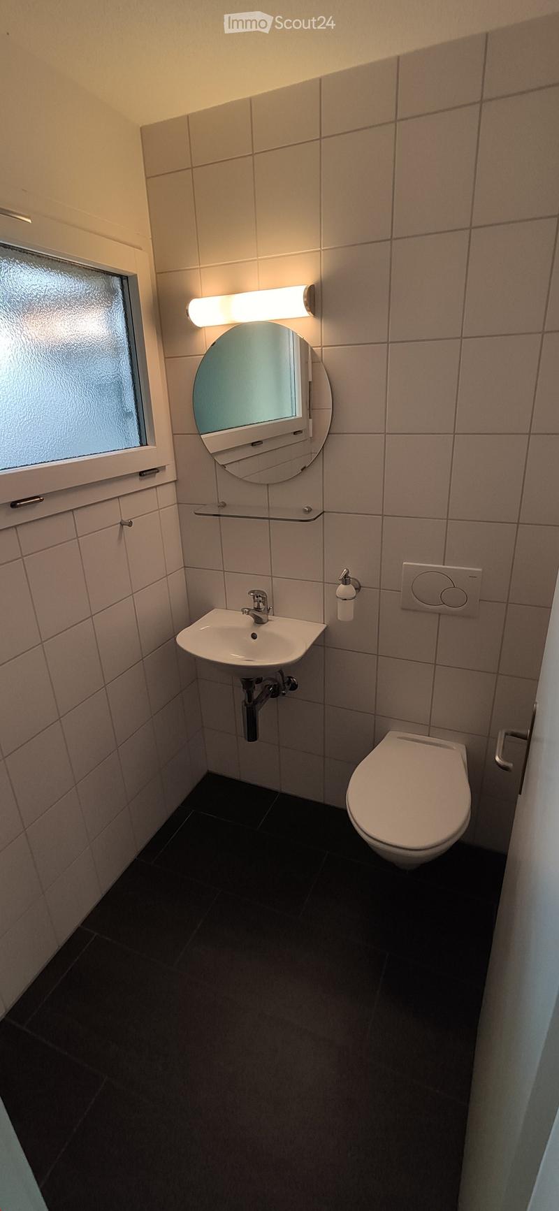 Modern bathroom with a white sink, toilet, round mirror with built-in lighting, and a frosted window. The walls are tiled in light beige, and the floor is dark.