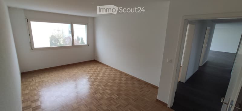 Empty room with wooden flooring and white walls, featuring large windows and an open doorway leading to another room.