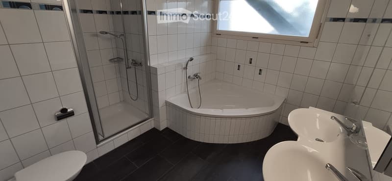 Modern bathroom with a corner bathtub, glass-enclosed shower, double sink, and toilet. The walls are tiled in white with black accents, and the floor is dark grey.
