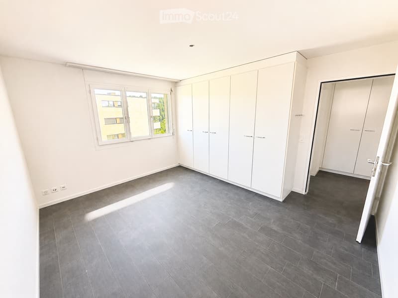 Empty room with white walls, grey tiled floor, built-in white wardrobes, and large windows letting in natural light.
